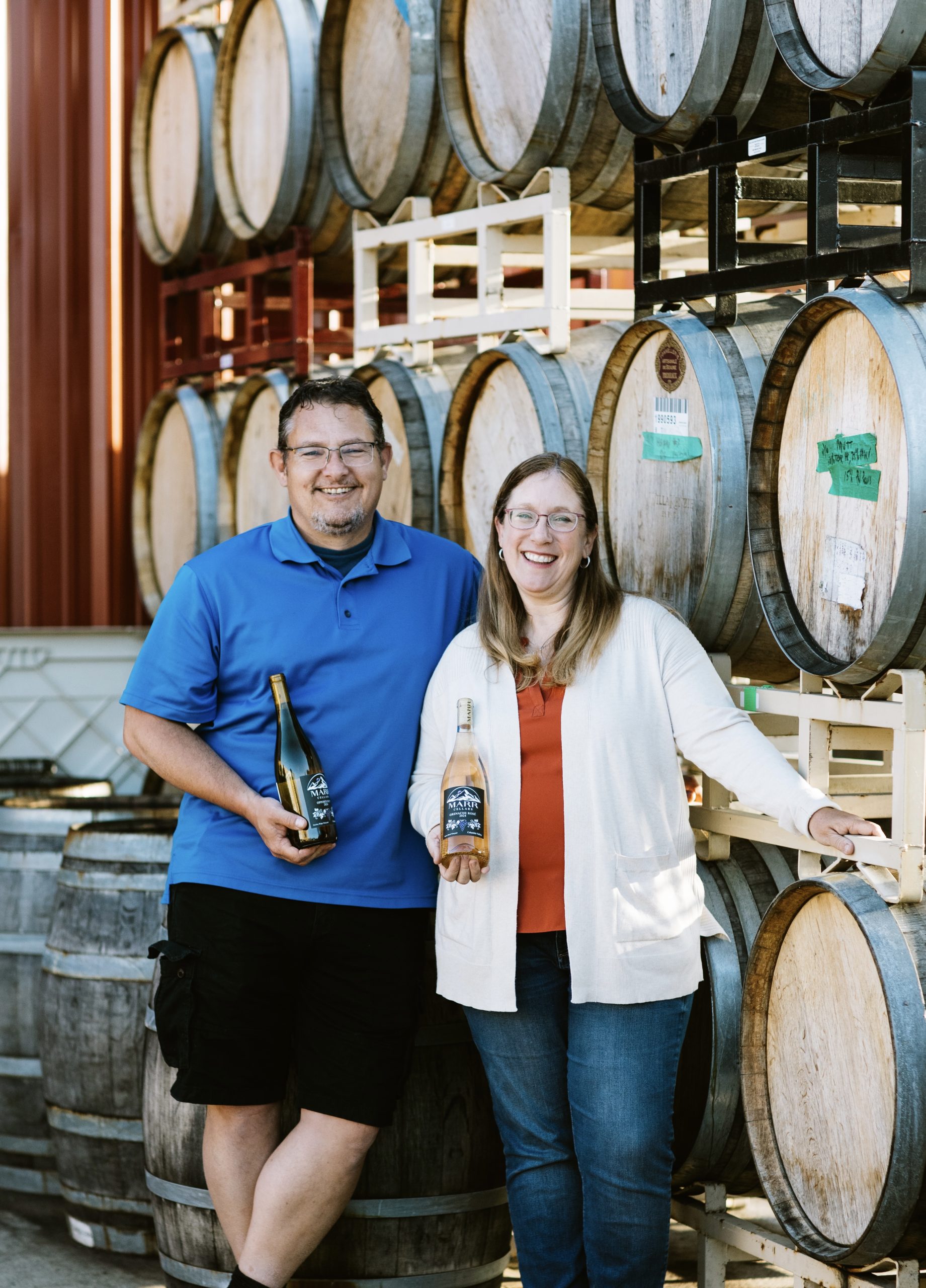 Two people posing with bottles of wine, in front of wine barrels