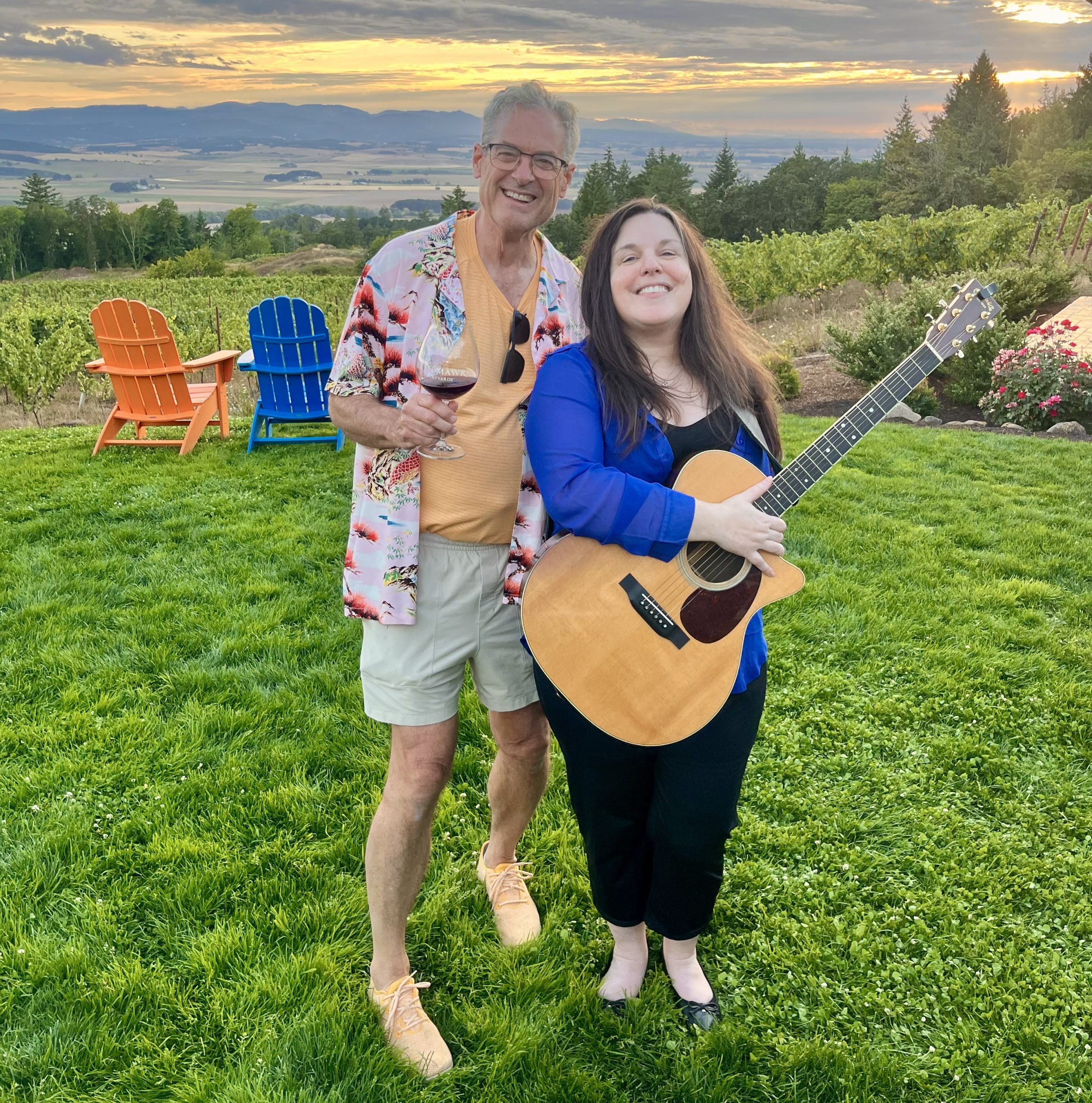 Two people in front of the vineyard, one holding an acoustic guitar